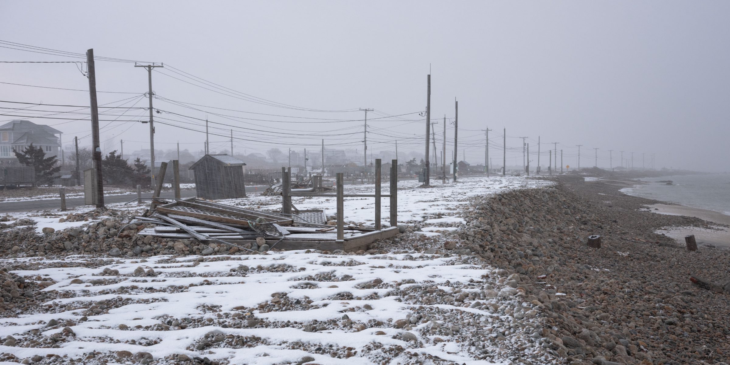 A snow-dusted rocky shoreline under heavy fog, a long row of utility poles and wires receding into gray, the sea at right.