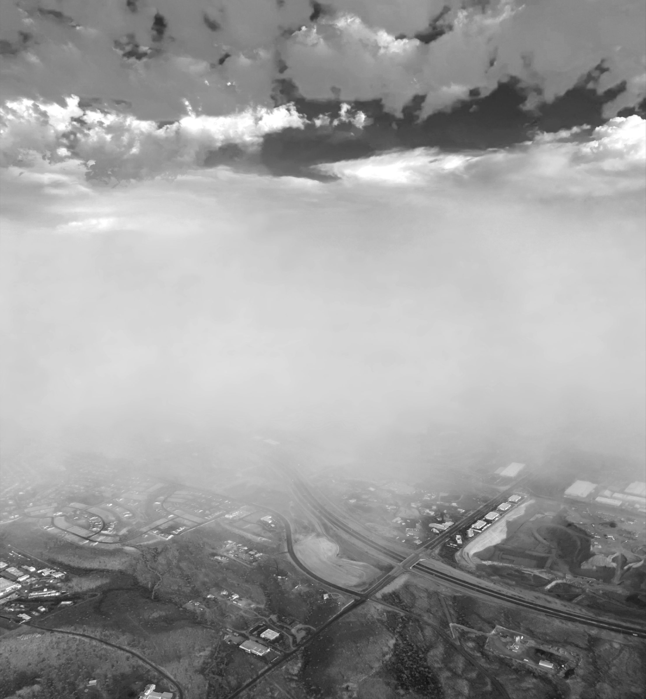 An aerial view from above the clouds looking down through fog onto a highway interchange and sprawl below.