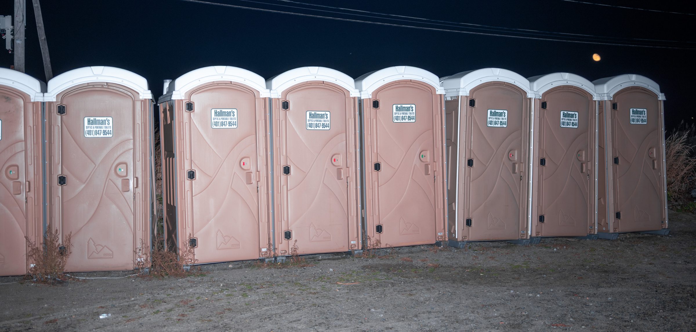 A row of seven pink portable toilets lined up at night against a black sky, a faint moon at right.