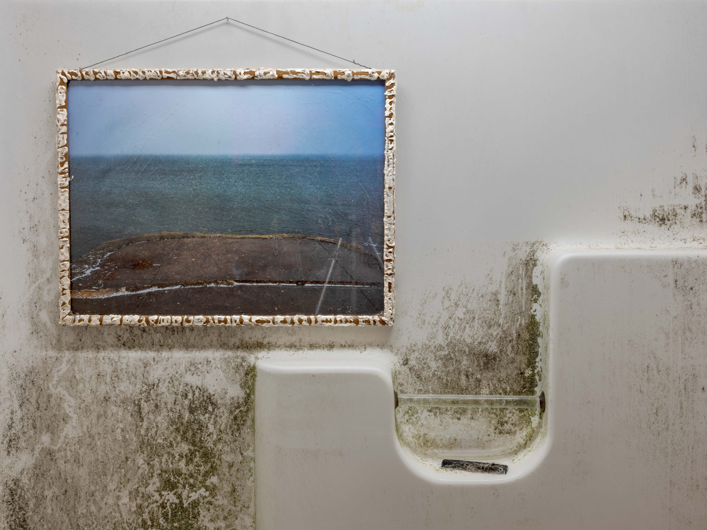 Installation view of 2nd hand art: a small seascape print in a caulk-built frame hung inside the grimy wall of a found tub-and-shower unit.