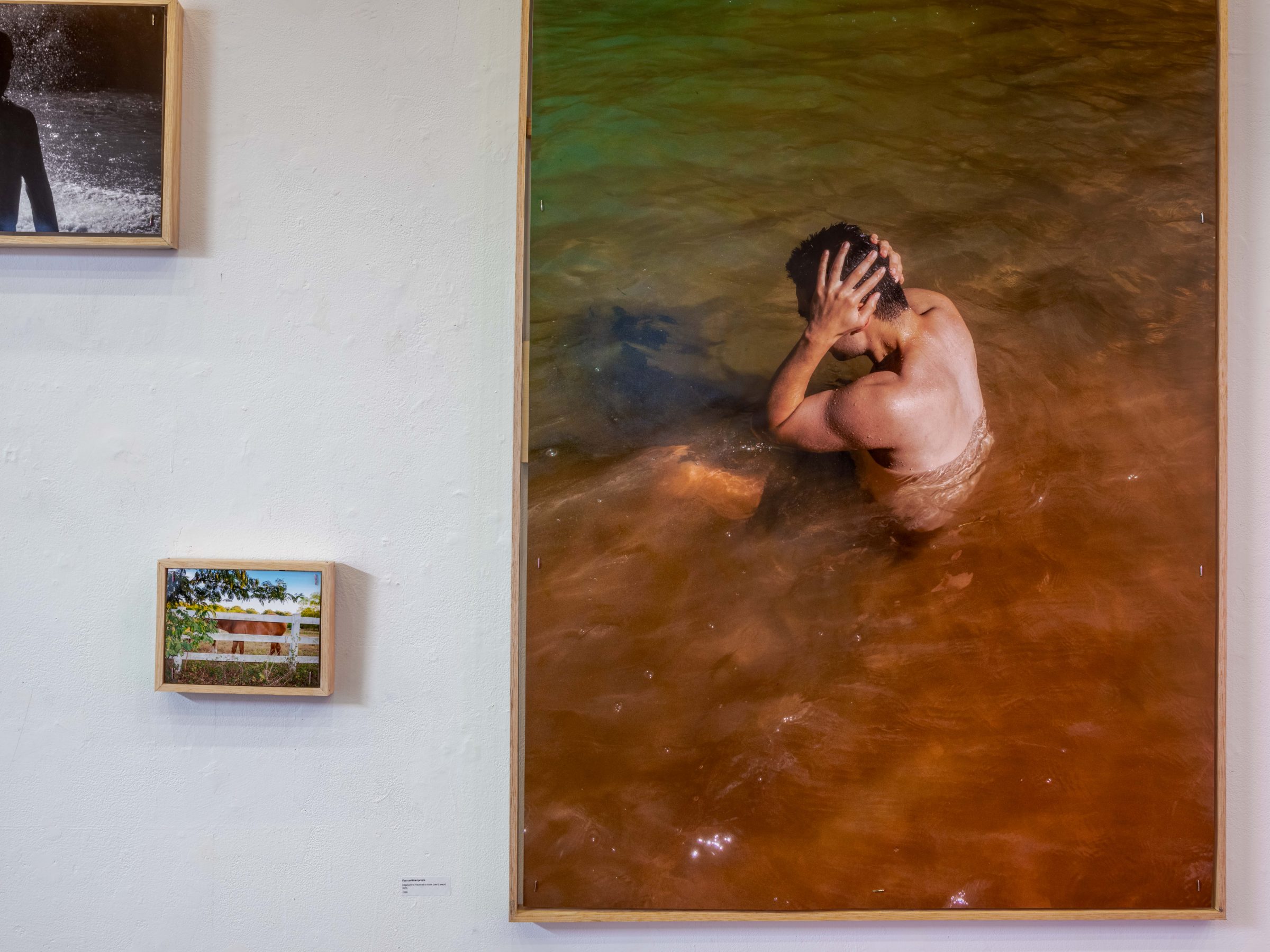 Installation view of 2nd hand art: the large vertical print of a figure bathing in orange water beside small framed prints of a silhouette and a horse.