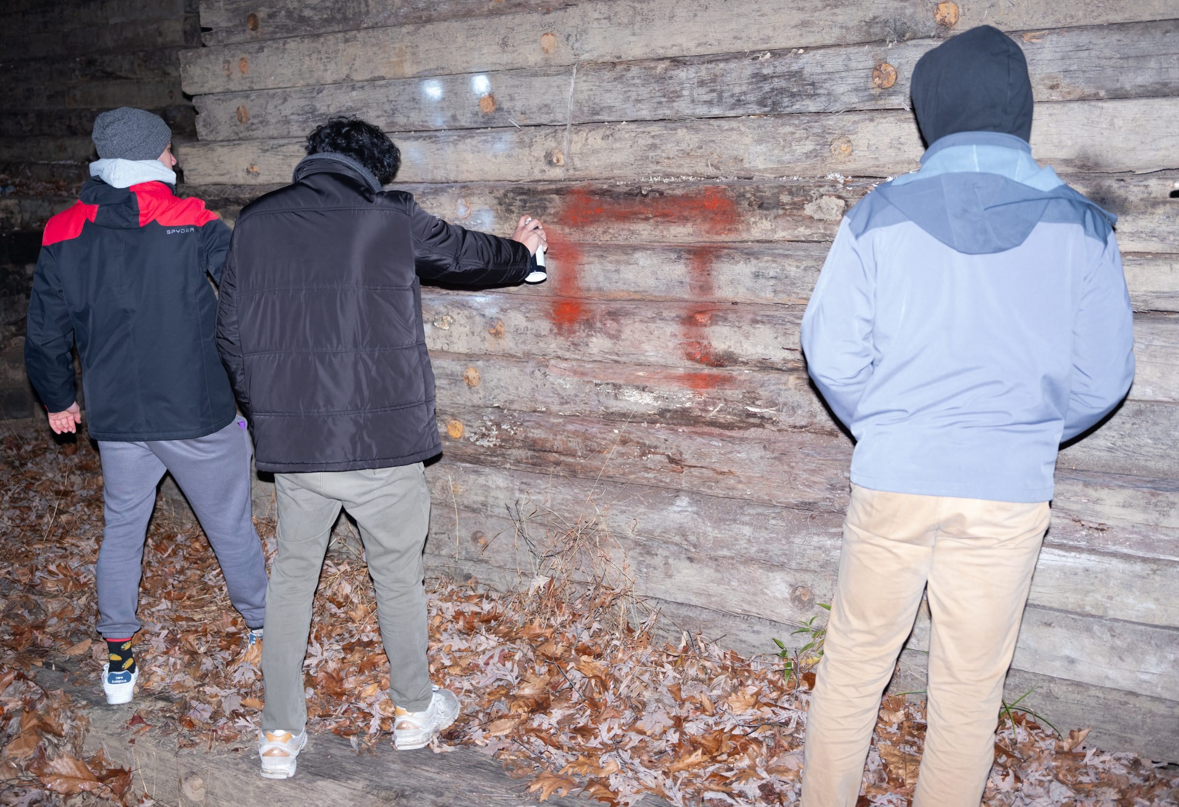 Three figures at night spray-painting a red mark onto the weathered log wall of a barn.