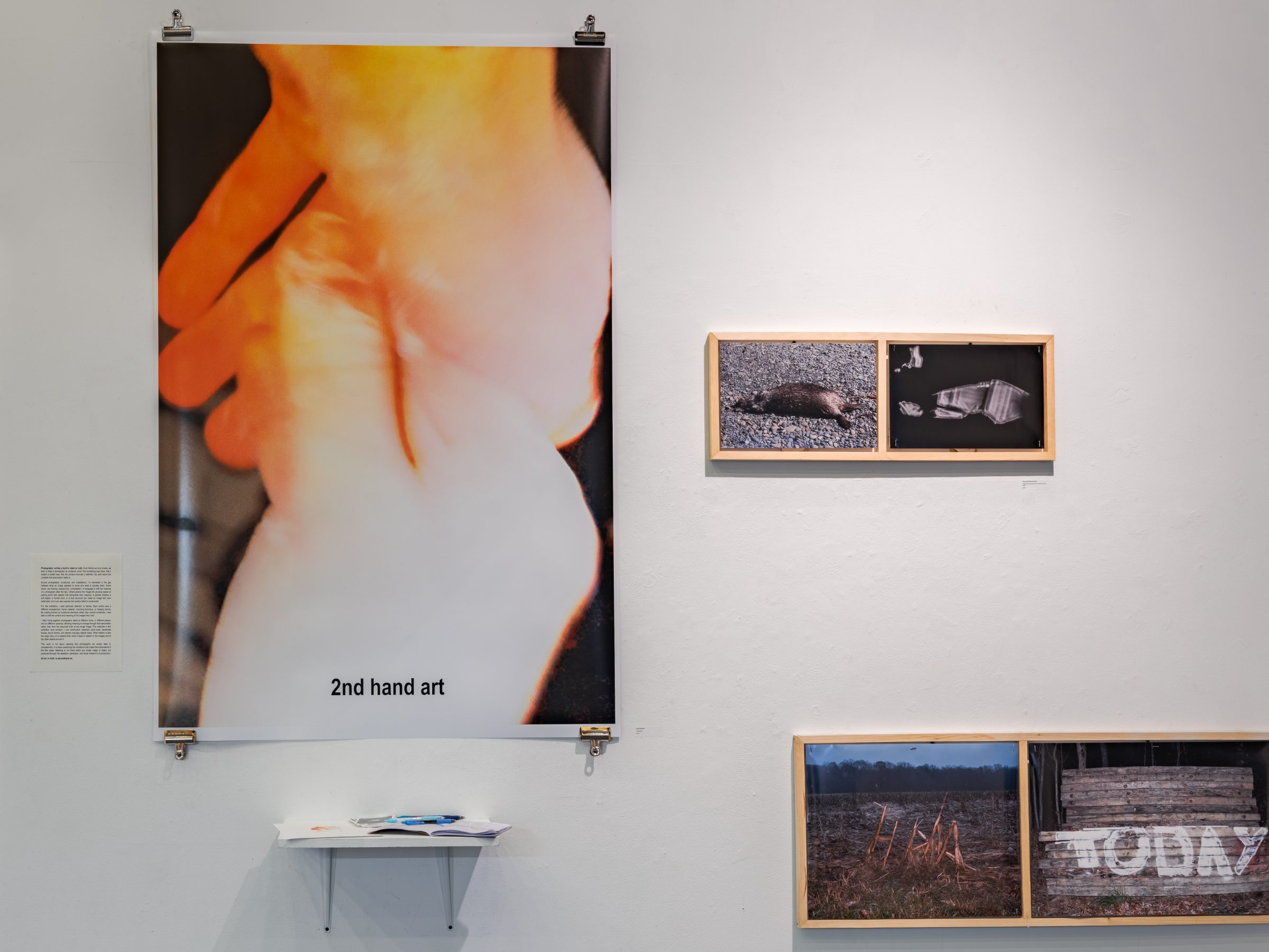 The large clipped poster of a backlit orange hand with the words '2nd hand art' across it, beside a wall text and two framed diptychs.