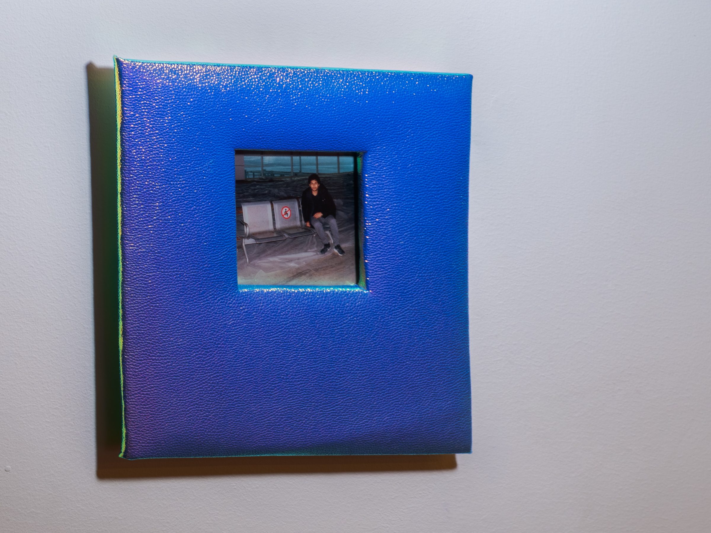 A young man seated on an airport bench, the snapshot inset into the iridescent blue-and-gold pebbled cover of an altered photo album, seen at a slight angle.