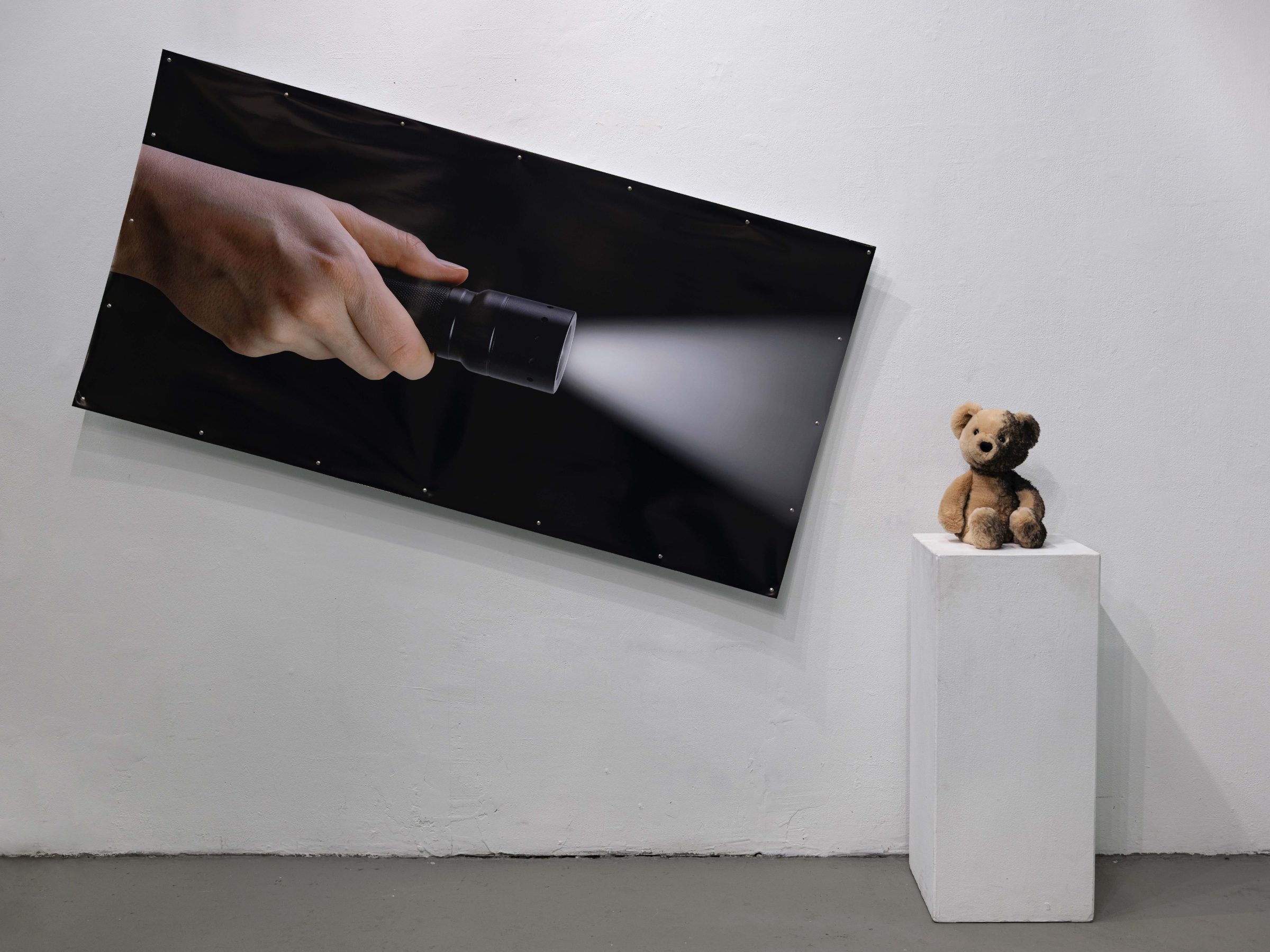 A wide black print of a hand holding a flashlight, its beam cutting across the dark image, hung beside a worn teddy bear on a white plinth whose body is half-darkened with paint like a cast shadow.