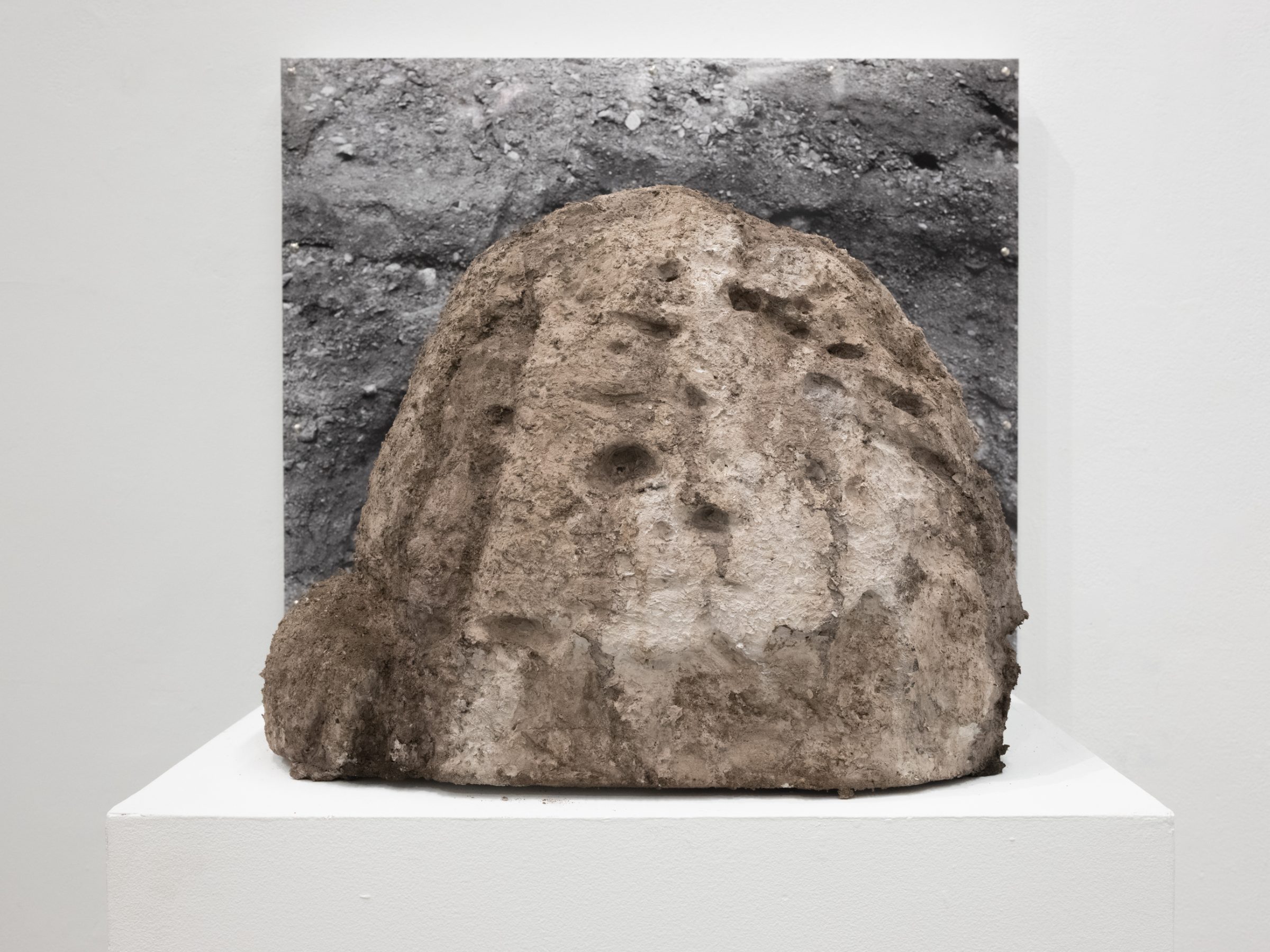 A cast sandy boulder sculpture on a white plinth, set in front of a grayscale photograph of a hole dug into rocky dirt.