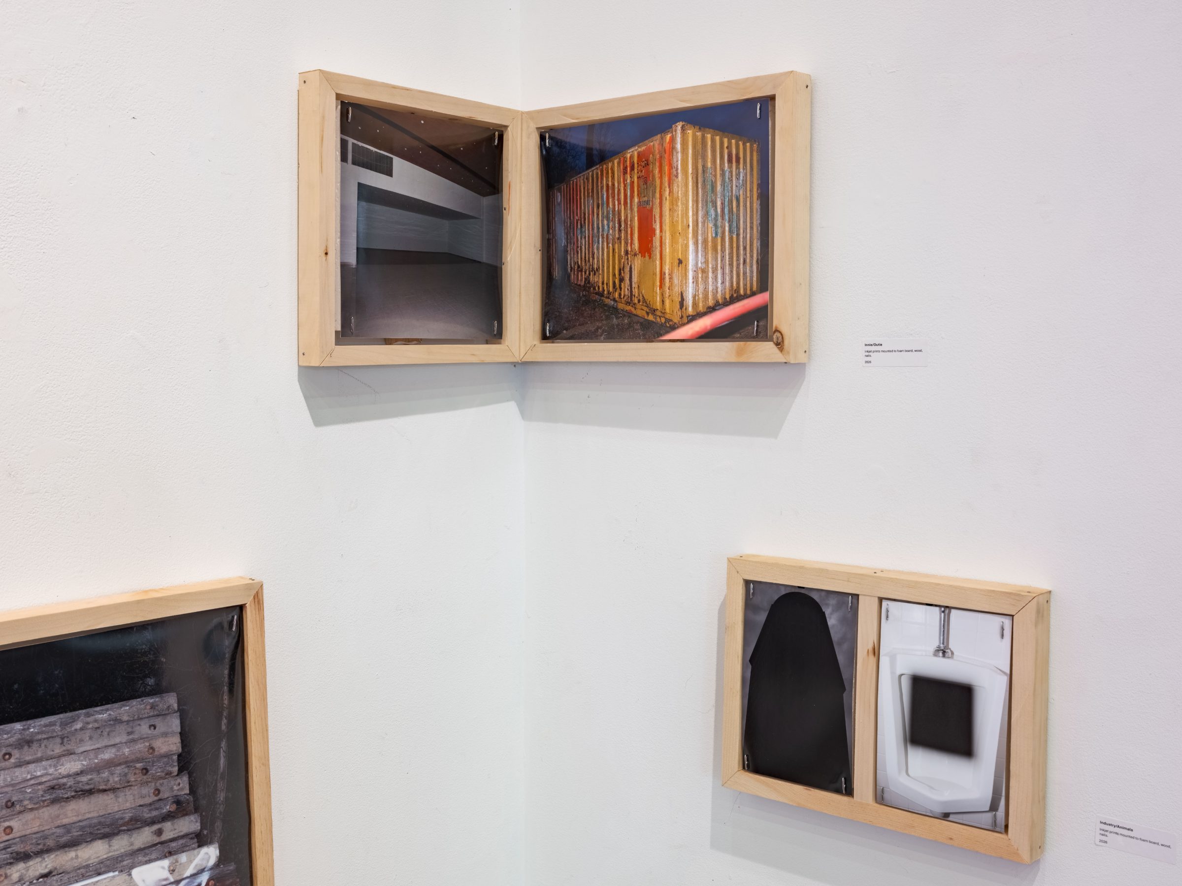 Two wood-box framed works wrapping a corner: an empty interior beside a rusted orange shipping container above, and a black silhouette beside a white urinal below.