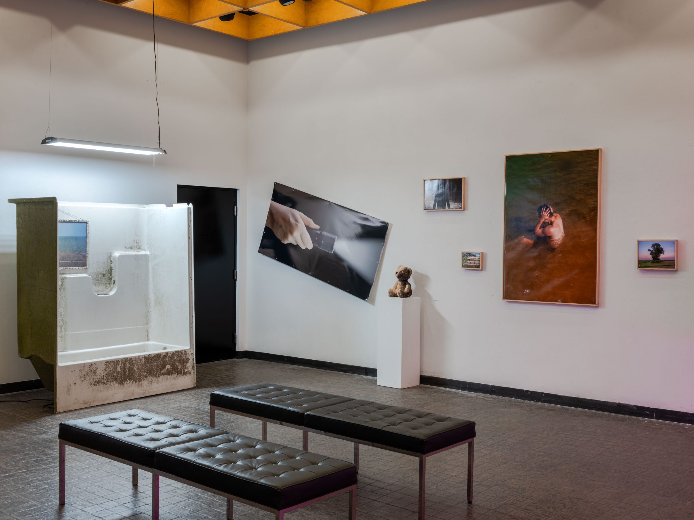 Installation view: a found tub-and-shower unit leaning against the wall, a tilted black print of a hand, a charred teddy bear on a plinth, and a large vertical print of a figure bathing in orange water.