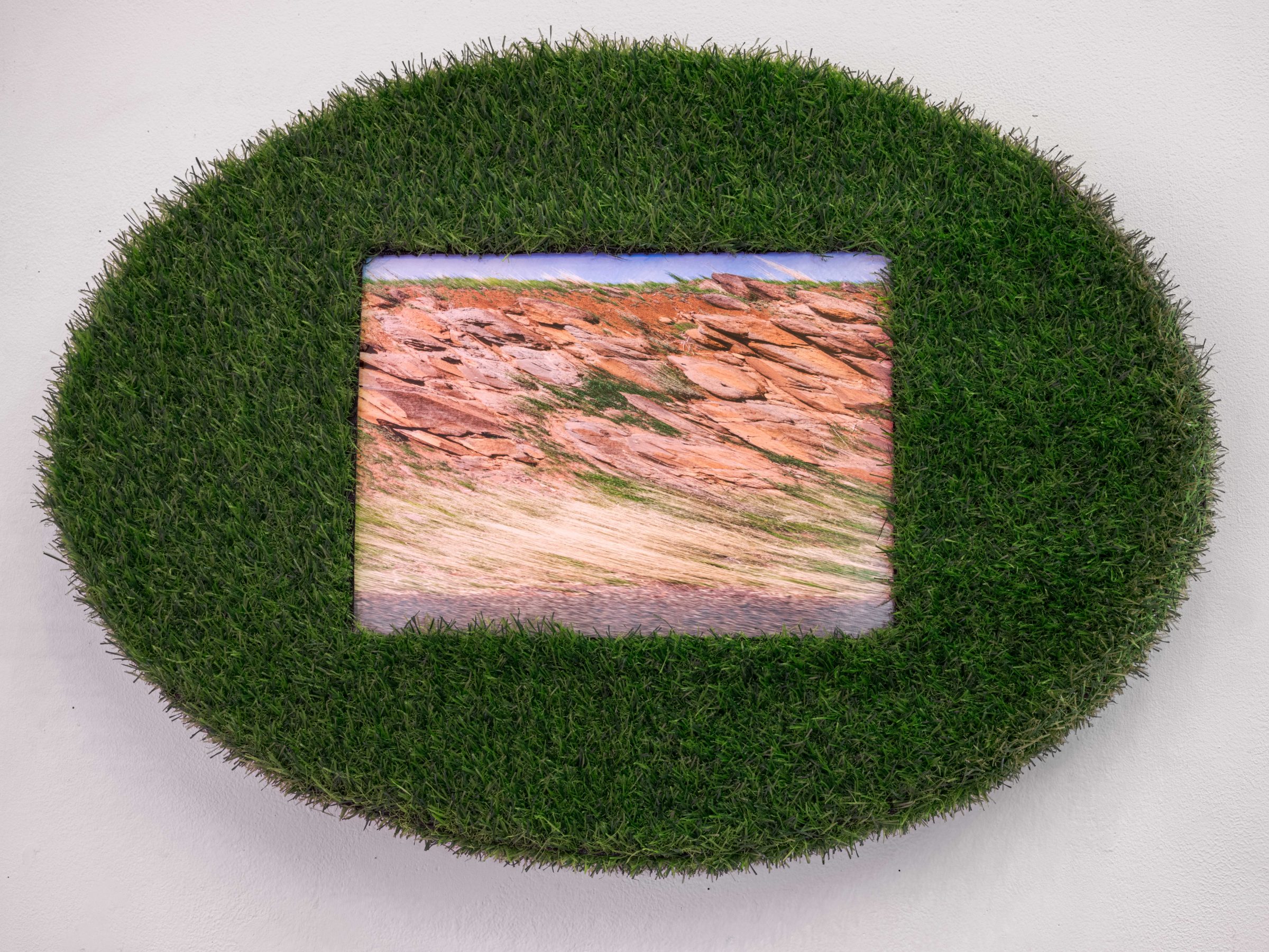 An oval cushion of artificial green turf mounted on the wall, framing a rectangular print of striated red-and-tan rock under a blue sky.