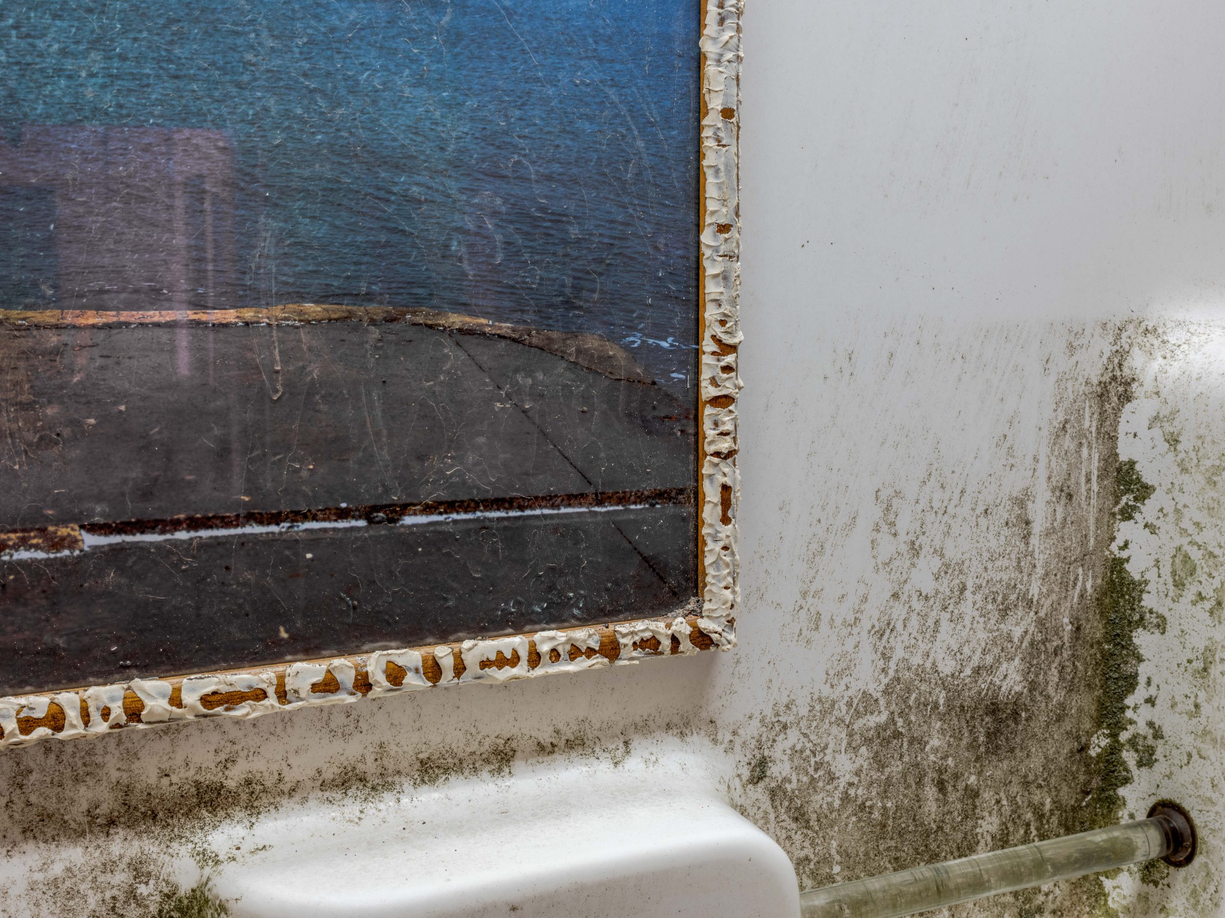 Detail: the corner of the seascape print, its edge thick with bathroom caulk, against the stained surface of the tub.