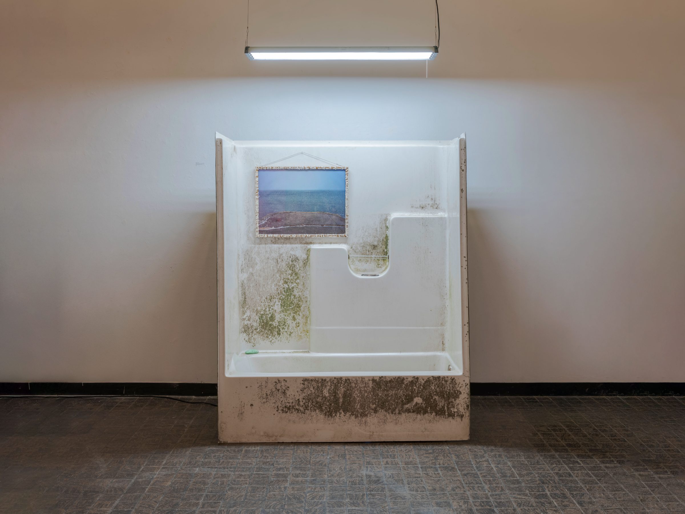A found tub-and-shower unit leaning against the gallery wall, mottled with grime, a small framed seascape hung inside it under a fluorescent light.