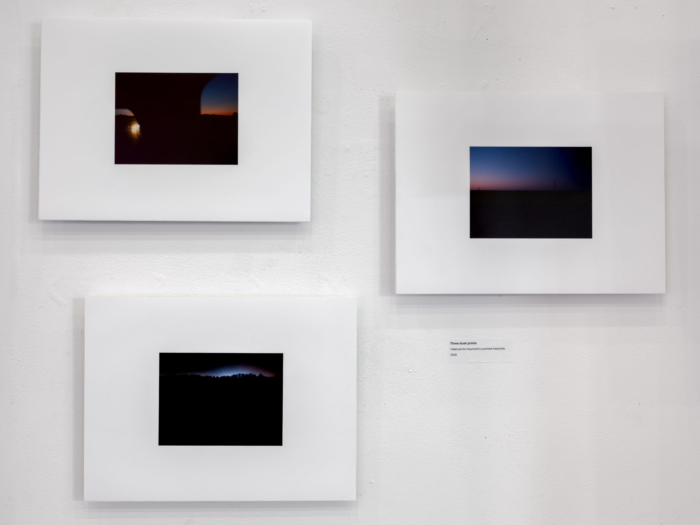 Three small dusk photographs mounted on white masonite panels and arranged across the wall: a window-lit horizon, a dim plain, and a dark treeline.